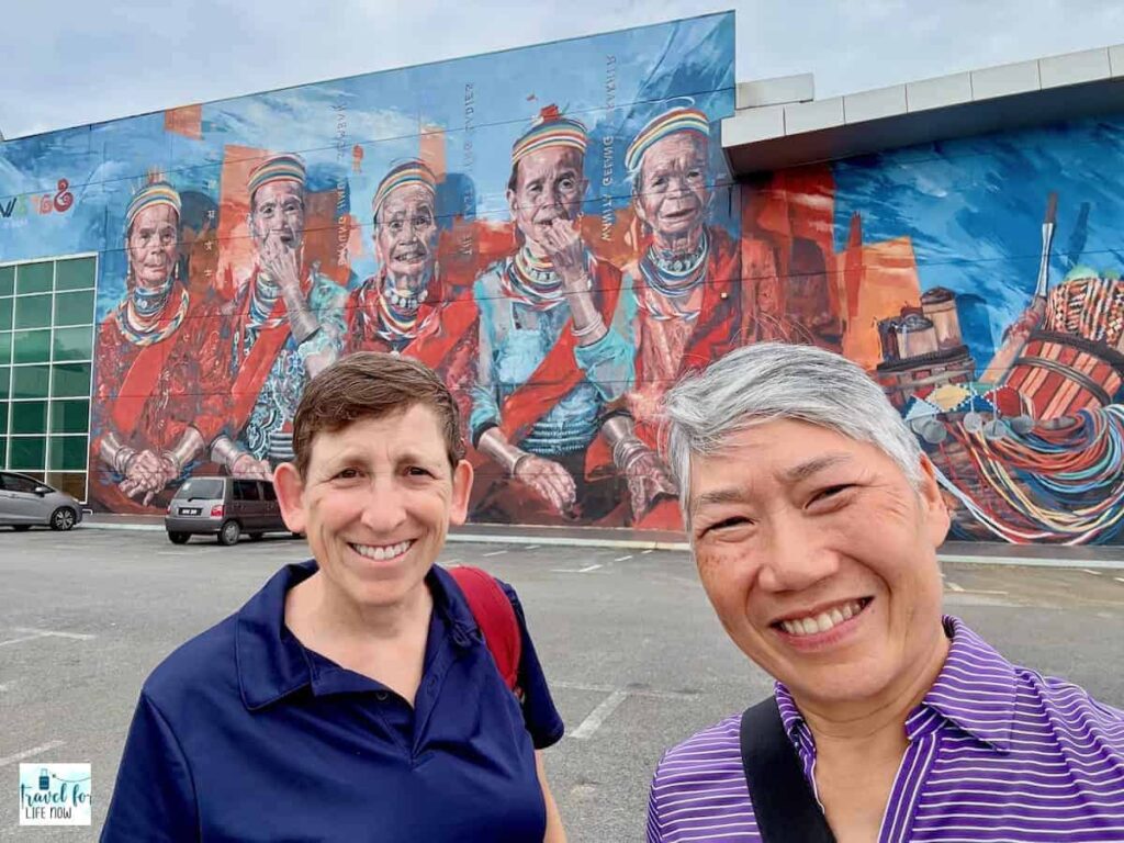 Sue and Reggie in front of a large mural of women in traditional attire.